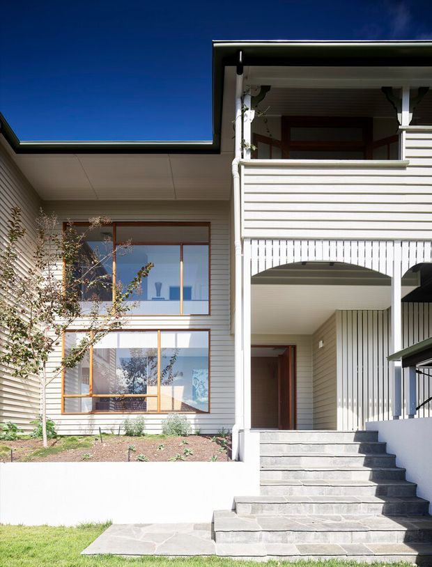 Front exterior view of a multi-level suburban house with a covered entry porch, broad grey stone steps, pale horizontal weatherboard cladding, timber-framed glazing, and a projecting upper balcony screened by horizontal and vertical slats. A small landscaped garden and lawn sit in front of the facade under clear daylight.