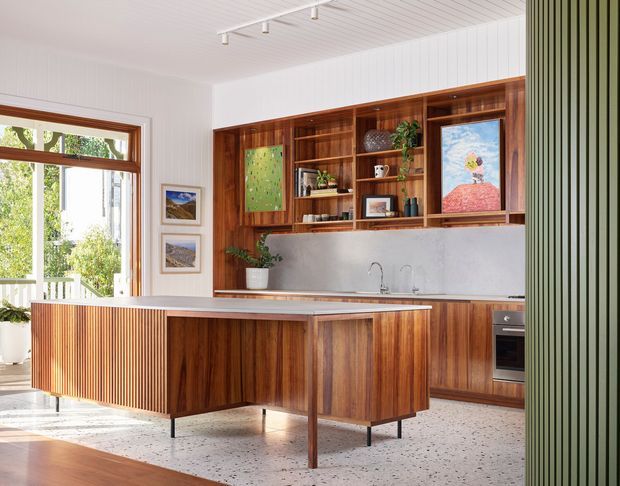Bright contemporary kitchen with a large timber island featuring vertical slatted detailing, extensive dark wood cabinetry, open display shelving, a pale stone or plaster splashback, and a terrazzo floor. White vertical wall panelling and ceiling boards add texture, while a large glazed door or window opens toward a leafy exterior. Artwork, plants, and shelving create a warm, curated mid-century-inspired interior.