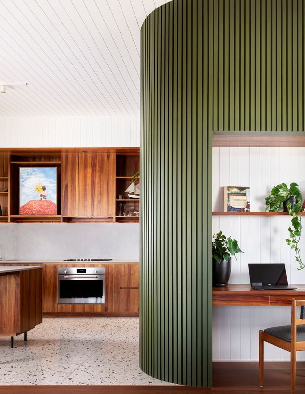 A bright, carefully detailed open-plan interior combining a warm timber kitchen with integrated cabinetry and oven, a terrazzo floor, and a compact study nook. A striking curved olive-green vertical-ribbed partition separates the spaces, while the study area features white vertical wall panelling, timber shelving and desk joinery, indoor plants, and a timber-framed chair. The overall composition is contemporary with strong mid-century-inspired timber detailing and a refined, warm palette.