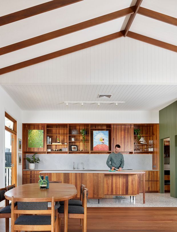 Bright open-plan kitchen and dining area with a dramatic vaulted ceiling. White vertical wall and ceiling panelling is framed by exposed dark timber rafters and beams. A full wall of warm timber built-in cabinetry incorporates open shelving, artwork displays, appliances and a long pale benchtop. A person is preparing food at the island, while a round timber dining table and upholstered timber chairs occupy the foreground. A deep green vertically panelled wall provides contrast at right, and large glazed doors/windows bring in daylight.
