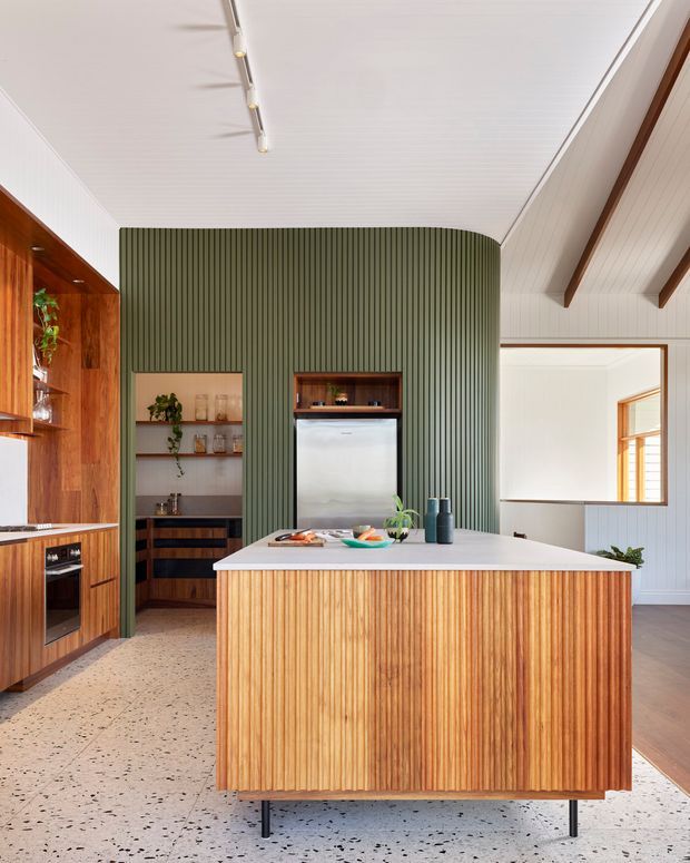A contemporary kitchen with a large central island featuring vertically fluted timber cabinetry and a pale benchtop. The room includes warm timber built-in cabinetry and appliances along the left wall, open shelving, a concealed refrigerator within a curved olive-green fluted feature wall, terrazzo flooring, and a white vertically panelled ceiling with exposed timber rafters. Linear track lighting and a large interior window complete the bright, warm space.