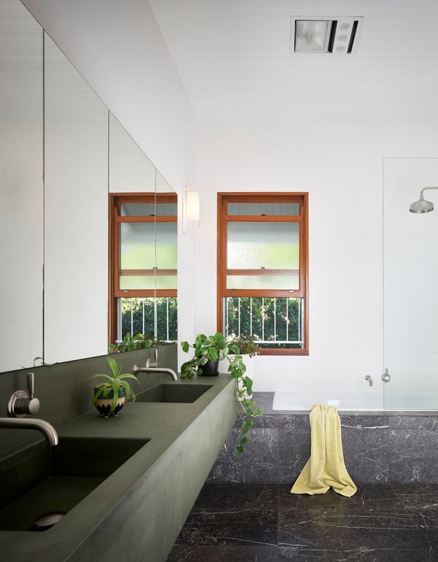 Bright contemporary bathroom with a long dark green double vanity, integrated basins, wall-mounted taps, and a large mirrored wall. Two timber-framed frosted windows sit above the vanity, while a freestanding white bathtub is set against a white wall and dark veined stone wall and floor finishes. Plants and a pale yellow towel add organic and warm accents.