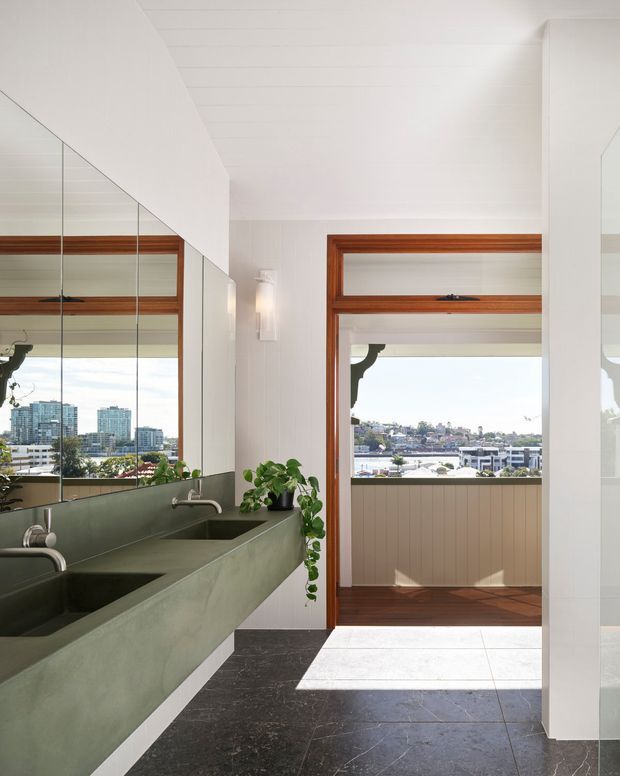 A bright, contemporary bathroom with a long green-toned double vanity, integrated basins and wall-mounted faucets. Large mirrored wall panels reflect the vanity and windows, while timber-framed windows and a glazed opening provide views across a waterfront or urban setting. The room has dark stone-look floor tiles, white vertical wall panelling, timber trim, and abundant natural light.