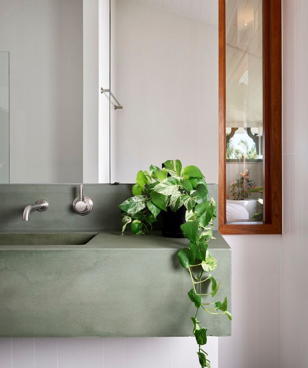 A close-up view of a contemporary bathroom vanity with a built-in muted green basin and backsplash, brushed metal wall-mounted tapware, and pale vertical wall tiles. A trailing variegated houseplant sits on the vanity, while a tall timber-framed mirror reflects part of the bright bathroom and greenery beyond.