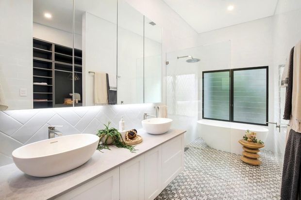 Bright contemporary bathroom with a freestanding oval bathtub, double vessel basins on a long white vanity, mirrored wall cabinets, a frameless glass shower, and a frosted-glass window. White geometric wall tiles and patterned encaustic-style floor tiles add visual interest, while integrated lighting creates a clean, airy atmosphere.