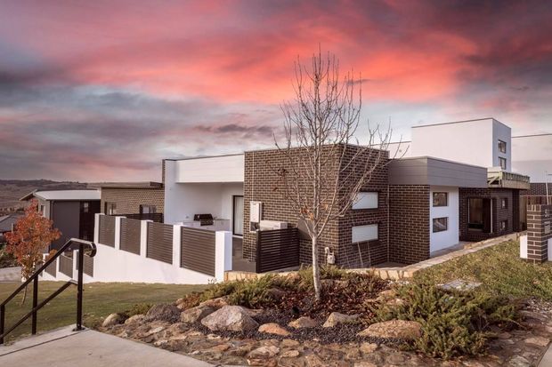 A contemporary single-storey residential house on a sloped suburban site, featuring dark brick masonry, white rendered and panelled volumes, horizontal flat roof forms, large rectangular windows, and a fenced front garden with exposed rock landscaping. A taller white cubic volume rises behind the main roofline. The image is an exterior architectural photograph at sunset.