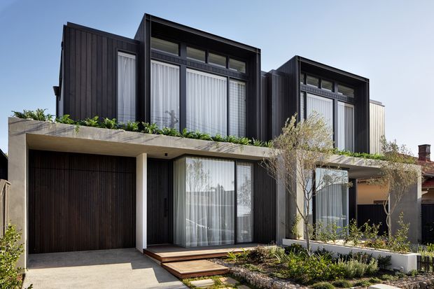 Exterior street-facing view of a contemporary multi-level residence with dark vertical cladding, projecting box-like upper volumes, extensive floor-to-ceiling glazing, a concrete terrace planter, integrated garage, and landscaped front garden.