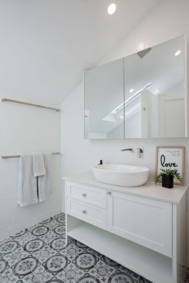 Bright, compact bathroom with a white wall-mounted vanity, integrated drawers and open lower shelf, white vessel basin, chrome tapware, large mirrored cabinet, white tiled walls, and decorative patterned floor tiles. A skylight reflected in the mirror adds natural light, while brushed-metal towel rails and a small framed print provide restrained accents.