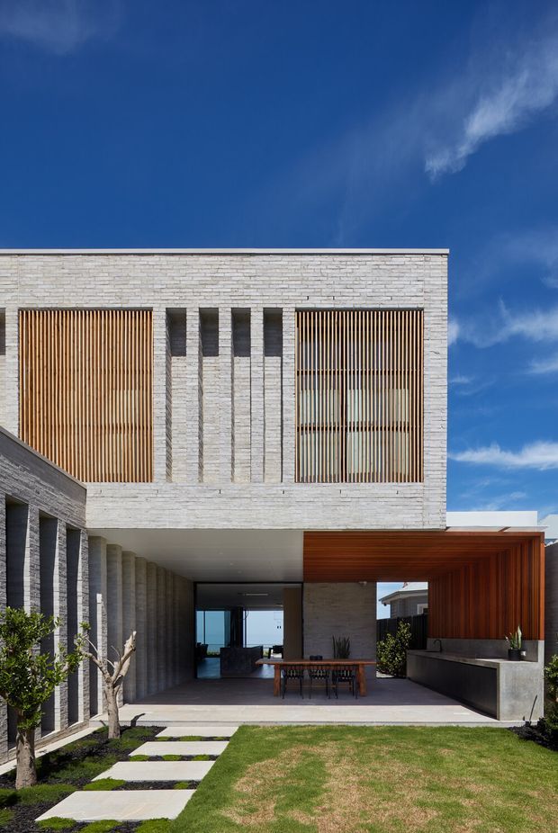 Contemporary two-storey residential facade with pale textured masonry, vertical timber screens, deep covered outdoor dining area, and a landscaped front lawn. The open ground-floor passage frames a glimpse of blue water or sky beyond, while strong geometric lines, recessed openings, and warm timber soffits contrast with the cool light masonry.