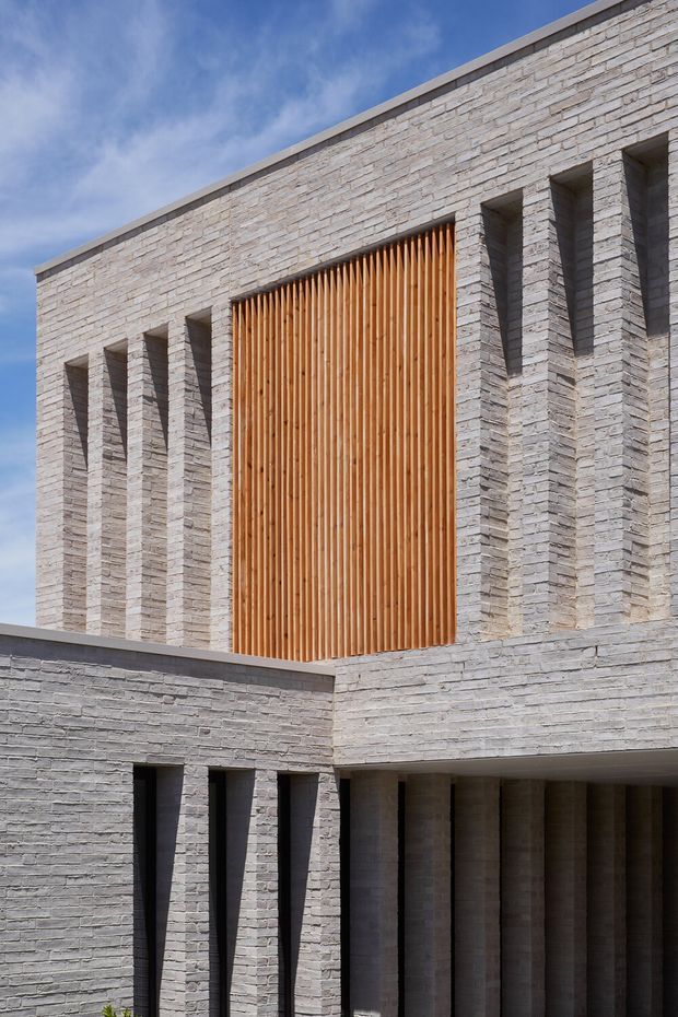 Exterior architectural detail of a contemporary masonry building. The facade is composed of pale grey stacked stone or brickwork with deep recessed vertical openings and projecting piers. A tall central opening is screened with closely spaced warm timber vertical slats, creating a strong contrast against the cool masonry. The flat roofline and minimalist geometric composition are clearly visible against the sky.