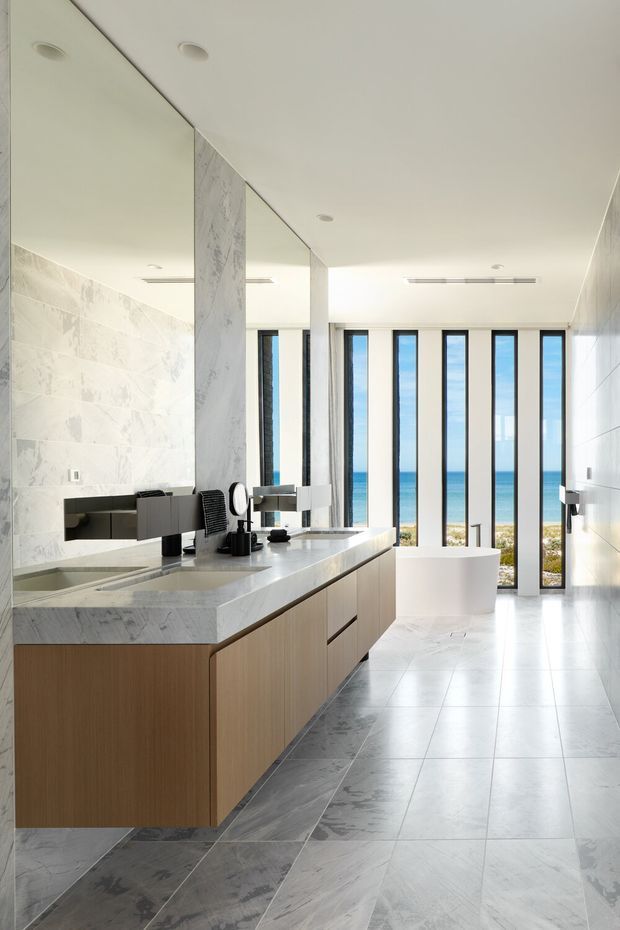 Bright, spacious contemporary bathroom with a large double vanity, integrated basins, wall-mounted dark fixtures, marble-look wall and floor tiles, and light timber cabinetry. A freestanding white bathtub sits beside full-height narrow windows overlooking a sandy coastal beach and ocean. The room has a clean, luxurious, highly refined feel with recessed ceiling lights and expansive glazing.