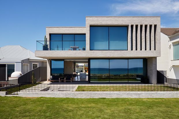 Front exterior view of a contemporary multi-level coastal residence with a rectilinear light-brick facade, extensive dark-framed glazing, projecting upper floor, vertical sun-shading fins, balcony, and a fenced lawn. Ocean views are visible through the ground-floor glazing and beneath a clear blue sky.