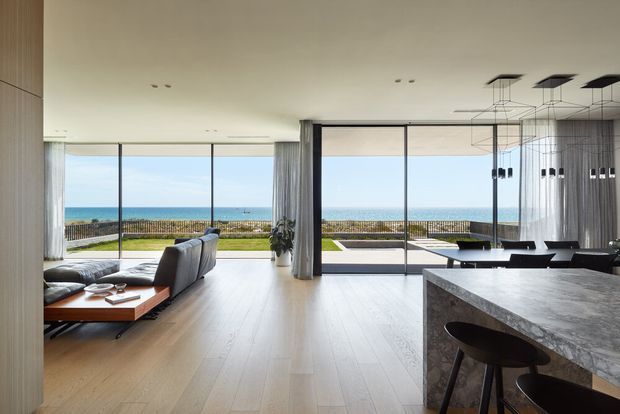 A bright, open-plan coastal interior with a living room, dining area, and kitchen opening onto a terrace and ocean views. Full-height sliding glass doors, pale timber flooring, neutral furnishings, a dark stone kitchen island, sheer curtains, and contemporary linear pendant lighting create a refined, spacious atmosphere.