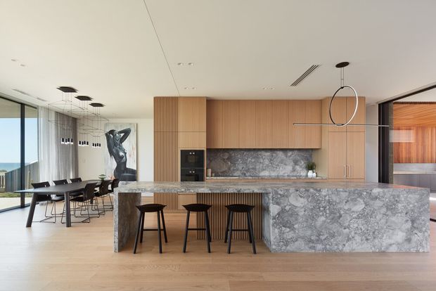 A refined open-plan kitchen and dining area with a large marble-look stone island and integrated waterfall end panel. Warm timber cabinetry and vertical timber battens contrast with the grey veined stone, while three dark stools sit at the island. The adjoining dining zone has a dark table and chairs beside floor-to-ceiling glazing, with contemporary pendant and linear lighting. A sculptural artwork and glimpses of an adjacent timber-lined room are visible in the background.
