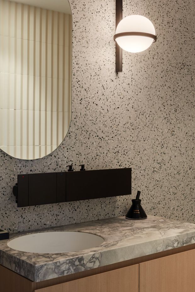 A refined contemporary bathroom vanity featuring a thick veined stone benchtop with an integrated oval basin, dark floating wall-mounted cabinetry, and warm timber cabinetry below. The walls are finished in a speckled terrazzo-like surface, complemented by a rounded-edge mirror with vertical ribbed detailing and a sculptural globe wall sconce in a dark metal finish.