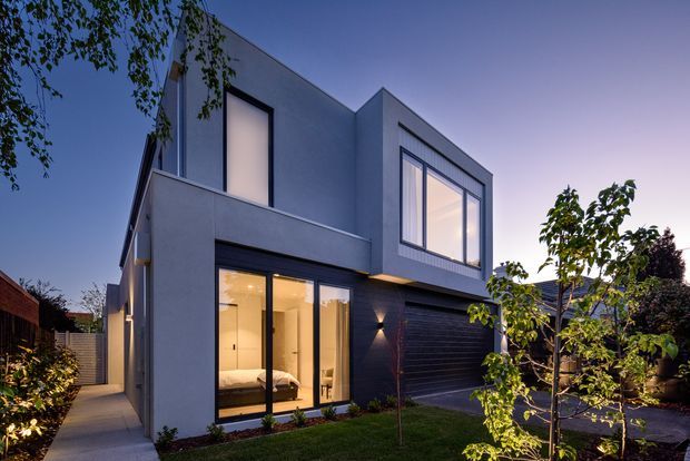 Twilight exterior view of a contemporary multi-level home with a clean geometric form, smooth light-grey rendered walls, dark horizontal cladding, large black-framed windows, and a dark garage door. Warm interior lighting glows through the glazing, with a narrow paved side path, lawn, and landscaped suburban garden in the foreground.