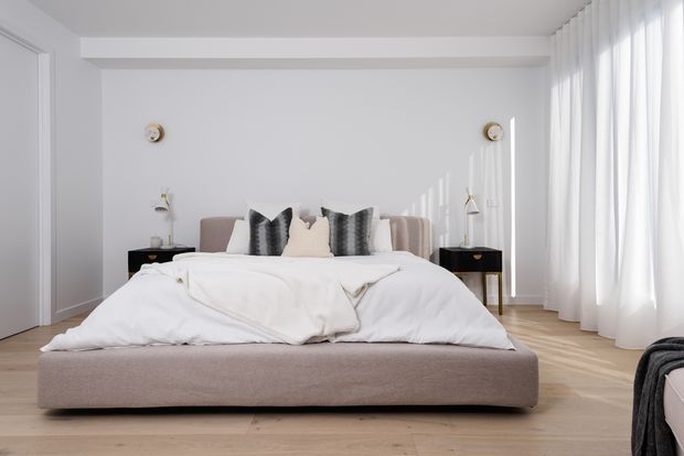Bright, refined bedroom with a low upholstered platform bed, white bedding, neutral decorative cushions, black bedside tables, wall-mounted reading lights, pale timber flooring, and full-height sheer curtains filtering abundant daylight. The room has a clean, contemporary hotel-like appearance with soft neutral tones.