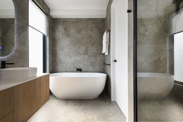 A contemporary bathroom featuring a freestanding white bathtub against grey stone-look wall and floor surfaces. A timber vanity with a white vessel basin sits beneath a rounded mirror, while a black-framed glass shower enclosure and reflected bath are visible on the right. The space has clean lines, restrained detailing and a neutral, tactile material palette.