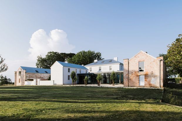 A broad exterior view of a group of low-rise buildings set behind an expansive lawn. The composition combines white-painted contemporary volumes with pitched metal roofs and skylights, a glazed ground-level connector, and a weathered exposed-brick masonry building. Mature trees frame the property, with a clear blue sky and open rural-looking grounds.