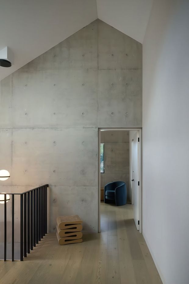 A minimalist interior landing or hallway with a tall, gabled wall clad in exposed concrete panels, visible form-tie marks, and neat panel joints. Pale timber flooring runs along the corridor, bordered by a black metal stair balustrade. A sculptural stacked timber stool sits against the concrete wall, while an open doorway reveals a small seating area with a dark blue armchair. The space is softly lit and vertically dramatic, with a restrained neutral palette and industrial-modern character.