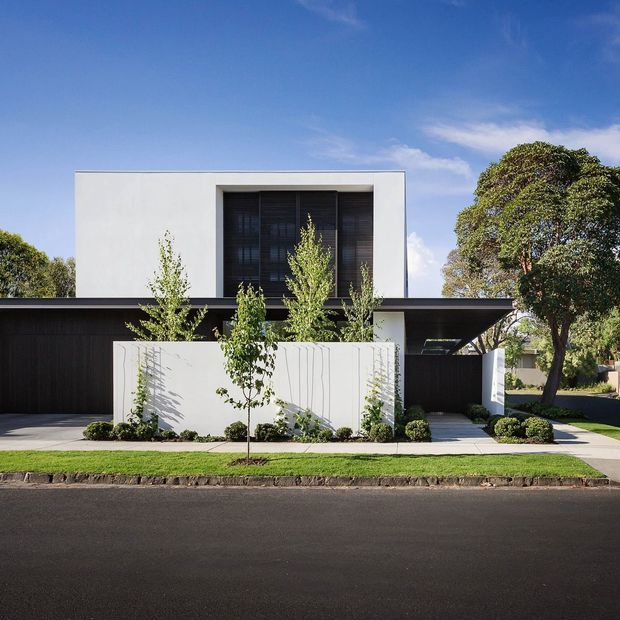 Street-facing contemporary residence with a crisp white two-storey upper volume, deep dark timber-clad ground level, recessed upper glazing with vertical screening, and a minimalist white boundary wall. Small landscaped planting beds, mature trees, a paved driveway and pedestrian path frame the facade.