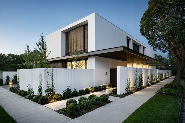 A contemporary two-storey residence with crisp white cubic volumes, deep flat roof overhangs, recessed glazing, and warm timber screening. The home is set behind white privacy walls with a landscaped front garden, clipped shrubs, climbing plants, paved paths, and subtle landscape lighting.