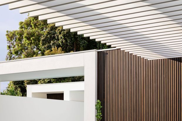 A close-up view of a contemporary building exterior featuring crisp white rendered walls, a large slatted pergola overhead, and a tall vertical timber screen forming a strong architectural feature. Mature greenery is visible behind the structure.