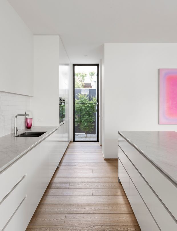 Bright, minimalist galley kitchen with white handleless cabinetry on both sides, pale stone benchtops, a stainless-steel sink and tap, and wide-plank timber flooring. A full-height glazed door at the end opens to a planted outdoor area, while a vivid pink artwork provides a strong accent on the otherwise white interior.