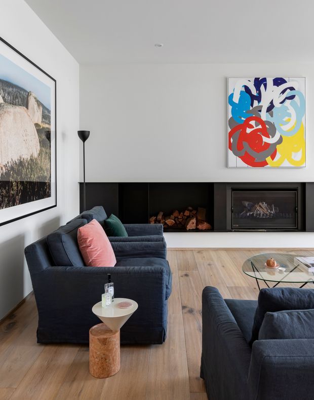 Contemporary living room with a low, built-in black fireplace wall and wood-burning firebox, pale timber flooring, white walls, and dark navy upholstered sofas and armchairs. A tall black floor lamp, sculptural side table, glass coffee table, large framed landscape photograph, and colourful abstract artwork add visual interest. The room has a clean, refined look with strong contrast and abundant natural light.