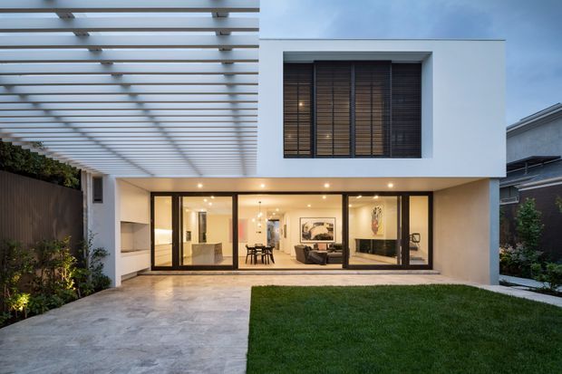 Contemporary two-storey residential home viewed from the rear courtyard, with a clean white rendered facade, extensive black-framed ground-floor glazing, upper-level timber screening, and a broad translucent pergola over the paved outdoor area. The illuminated interior reveals an open-plan living and dining space, while a lawn and planted boundary soften the modern architecture.