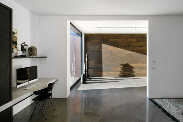 A bright, minimalist modern interior viewed from a living area toward an adjoining open space. White walls and ceiling frame a dark polished floor, a compact wall-mounted television and floating shelf, and a small black desk chair. The rear space features a dramatic dark timber feature wall, tall glazing, strong angular sunlight, and sculptural black decorative objects. A partial rug and dark built-in joinery are visible in the foreground.