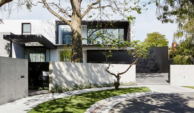 Contemporary two-storey residential facade viewed from the street, featuring crisp white rendered walls, dark vertical cladding, black-framed glazing, projecting flat roof forms, and a minimalist concrete boundary wall and driveway. Mature trees and restrained landscaping soften the geometric architecture.