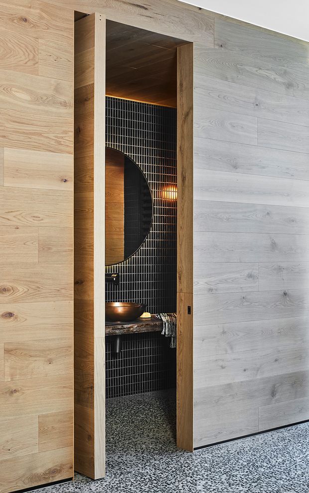 A narrow, refined bathroom viewed through an open timber-framed doorway. The room features dark vertical grid-pattern wall tiles, a round black-framed mirror, a vessel basin on a dark stone benchtop, warm concealed and pendant lighting, and a speckled terrazzo floor. Pale timber wall panelling surrounds the doorway and continues across the adjacent wall, creating a strong contrast with the dark bathroom interior.