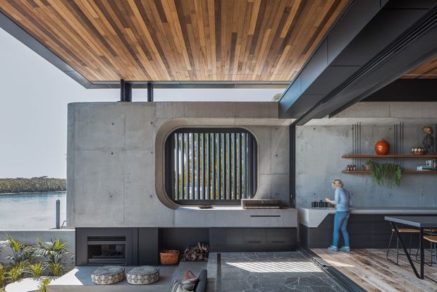 A contemporary open-plan kitchen and dining space with a strong indoor-outdoor connection. The room features a concrete wall and built-in kitchen joinery, a dark stone benchtop, a rounded rectangular window with vertical screening, exposed timber ceiling boards, dark steel structural framing, and a dining table opening toward a waterfront view. A built-in fireplace or hearth is visible below the kitchen counter, with shelving and minimal decor along the rear wall.