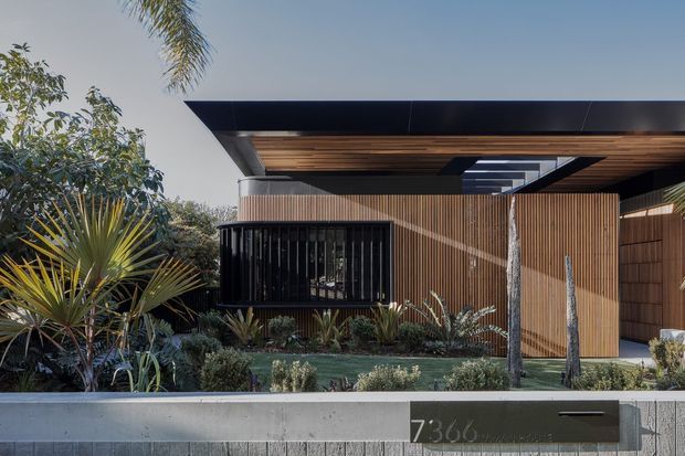 A contemporary single-storey house facade viewed from the street, with a broad flat projecting roof, timber-lined soffit, dark metal roof edges, extensive vertical timber screening, dark-framed glazing, and a landscaped front garden behind a low concrete boundary wall. A skylight or roof opening is visible beneath the covered entry/carport-like overhang.
