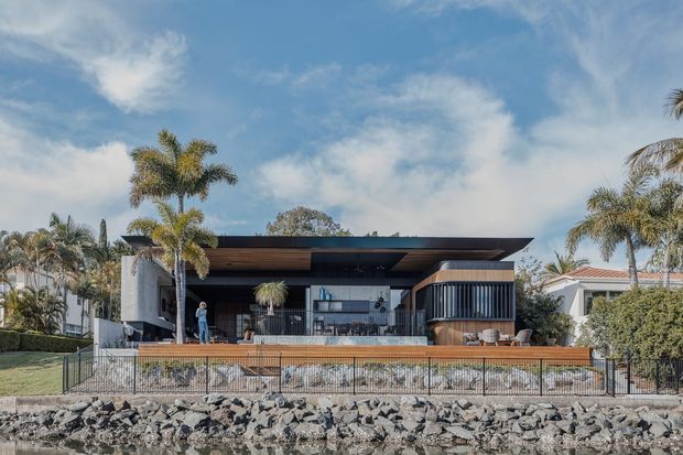 A contemporary single-storey waterfront residence viewed from across a rocky shoreline. The home features a broad low-pitched roof with deep overhangs, dark horizontal cladding, extensive glazing, a timber-lined covered outdoor living area, and a raised timber deck with a glass and metal balustrade. Palm trees and neighbouring homes surround the property.
