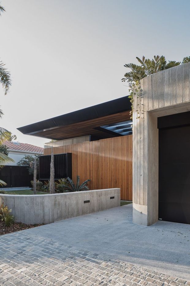 Exterior view of a contemporary residential entry and garage. The composition features a flat projecting roof with timber soffit, vertical timber screening, board-formed concrete walls and planter, a dark garage door, and a paved driveway. Tropical planting and a neighbouring tiled roof are visible in the background.
