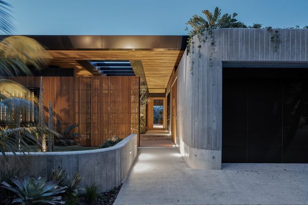 Contemporary residential entrance viewed from outside, featuring a sculptural concrete garage volume, timber-lined covered walkway, vertical timber screening, a skylight, and integrated landscaped planter beds. The composition uses strong horizontal and vertical forms with warm timber contrasting against cool grey concrete, photographed in soft evening light.