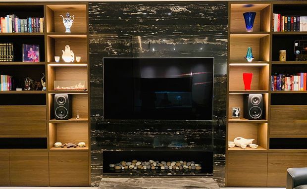 A dark, luxurious living room media wall with a centrally mounted television above a linear pebble-filled fireplace, framed by symmetrical timber built-in shelving and cabinetry. The shelving displays books, ceramics, glassware, and speakers, while a dramatic black stone feature wall creates a strong focal point.