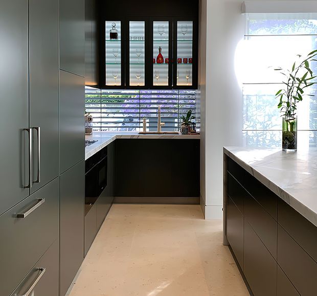 A contemporary galley-style kitchen with dark built-in cabinetry, long vertical handles, pale stone benchtops, and a matching central island or peninsula. The rear cabinetry includes a sink beneath a horizontal louvre window, while black-framed glass-front overhead cabinets display glassware and bottles. A large window provides abundant natural light, with a tall indoor plant placed on the island.