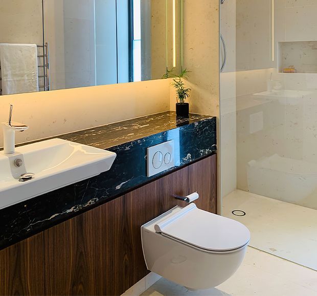 Contemporary bathroom featuring a wall-hung toilet, white vessel-style basin, dark veined stone vanity benchtop, vertically grained timber cabinetry, large mirror, and a frameless glass shower enclosure. Warm integrated lighting and a small plant add a refined, spa-like feel.