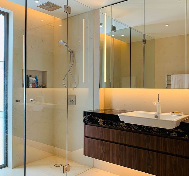 Contemporary bathroom with a frameless glass walk-in shower, pale stone-look wall and floor finishes, recessed shower niche, wall-mounted timber vanity, black marble-look benchtop, integrated white basin, and large mirrored wall cabinetry. Warm concealed lighting creates an inviting atmosphere.