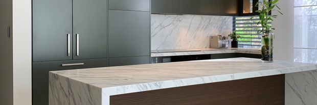Contemporary kitchen interior featuring a large marble-look island with a waterfall edge, dark green-grey full-height cabinetry, integrated appliances, and a matching marble-look splashback. Timber-toned island paneling, black-framed window glazing, and indoor greenery add contrast.
