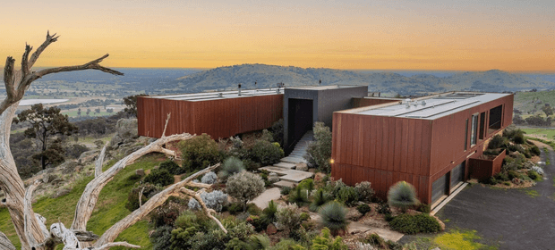 A contemporary low-profile residence composed of several rust-red rectangular volumes with flat roofs, set into a rocky, elevated landscape. Dark-framed glazing and a recessed central entry connect the forms, while native-style planting, exposed rock, and mature pale tree trunks surround the building. A broad valley and distant hills are visible beyond the site under warm evening light.