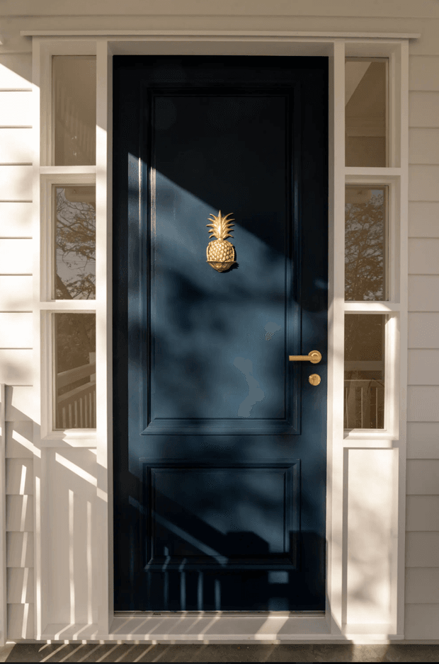 Front-facing exterior view of a traditional-style entry door. A dark navy-black raised-panel door with a decorative brass-gold pineapple knocker and matching brass hardware is framed by white trim and narrow sidelights. White horizontal weatherboard cladding surrounds the entry, with strong dappled sunlight and tree shadows across the facade.