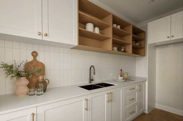 A bright, refined kitchen with white shaker-style cabinetry, a pale stone benchtop, white square-tile splashback, brass hardware, and a black undermount sink with a brushed metal tap. Open natural timber shelving provides warmth above the benchtop, while decorative cutting boards, plants, and ceramics add understated styling. The kitchen opens toward a hallway or adjacent space at right and has a warm timber floor.
