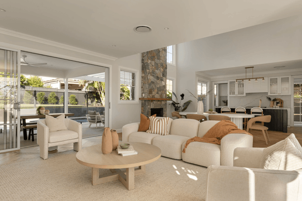 Bright open-plan living room with a large cream sectional sofa, upholstered armchairs, round timber coffee table, and neutral textured carpet. A tall rustic stone-clad fireplace forms a feature wall, while full-height glazed doors open to an outdoor terrace. The adjoining kitchen has white cabinetry, a light benchtop, dark lower cabinetry, and an island with timber stools. High-level glazing and an open connection between living, dining, and kitchen areas create an airy, contemporary interior with warm natural accents.
