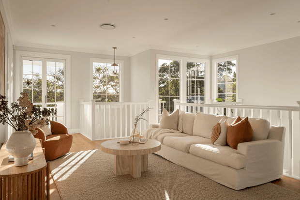 Bright, airy living room with a large cream sofa, rust-toned accent cushions and armchair, round timber coffee table, woven rug, white painted balustrade, and abundant multi-pane windows. Natural sunlight fills the room, with a neutral warm palette and relaxed contemporary styling.