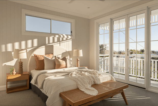 Bright, warmly styled coastal bedroom with a low-profile bed dressed in layered white and cream bedding, textured throw, and neutral cushions. The room has light vertical wall panelling, white-trimmed windows and full-height glazed French doors opening to a balcony or terrace. Natural sunlight creates strong linear shadows across the walls and floor, while timber bedside furniture, lamps, and a woven timber bench add warmth.