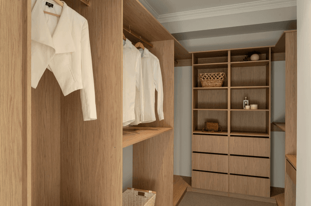 A warm, custom walk-in wardrobe with full-height wood-grain shelving, hanging rails displaying white shirts, open storage compartments, drawers, and integrated dressing-area joinery. The space has muted blue-grey walls, a carpeted floor, and a recessed ceiling with simple white cornices. Styling is practical and refined, with woven baskets and minimal accessories.